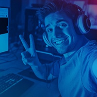Zdjęcie w kolorze niebieskim, młody człowiek ze słuchawkami siedzi przy biurku z komputerem i robi znak Zwycięstwa w kierunku kamery