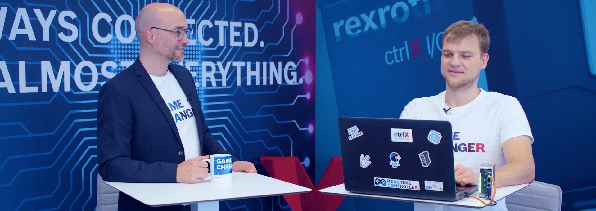 Gospodarz Christian Zentraf i jego gość Michael Schäfer stoją przy stołach i rozmawiają o programowaniu skryptów Pythona bezpośrednio na ctrlX CORE, sprzęcie sterującym rozwiązania automatyzacji ctrlX AUTOMATION.