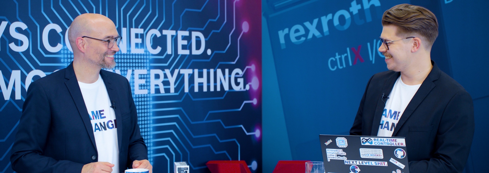 Gospodarz Christian Zentraf i jego gość Benedikt Rüb stoją przy stołach i rozmawiają o ctrlX Configurator, narzędziu konfiguracyjnym rozwiązania automatyzacji ctrlX AUTOMATION.