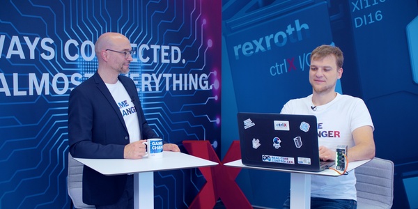 Gospodarz Christian Zentraf i jego gość Michael Schäfer stoją przy stołach i rozmawiają o programowaniu skryptów Pythona bezpośrednio na ctrlX CORE, sprzęcie sterującym rozwiązania automatyzacji ctrlX AUTOMATION.