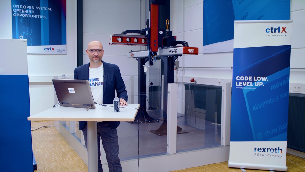 Host Christian Zentraf stands at a meeting table and shows in a live demo how the automation solution ctrlX AUTOMATION controls and drives a pick-and-place application