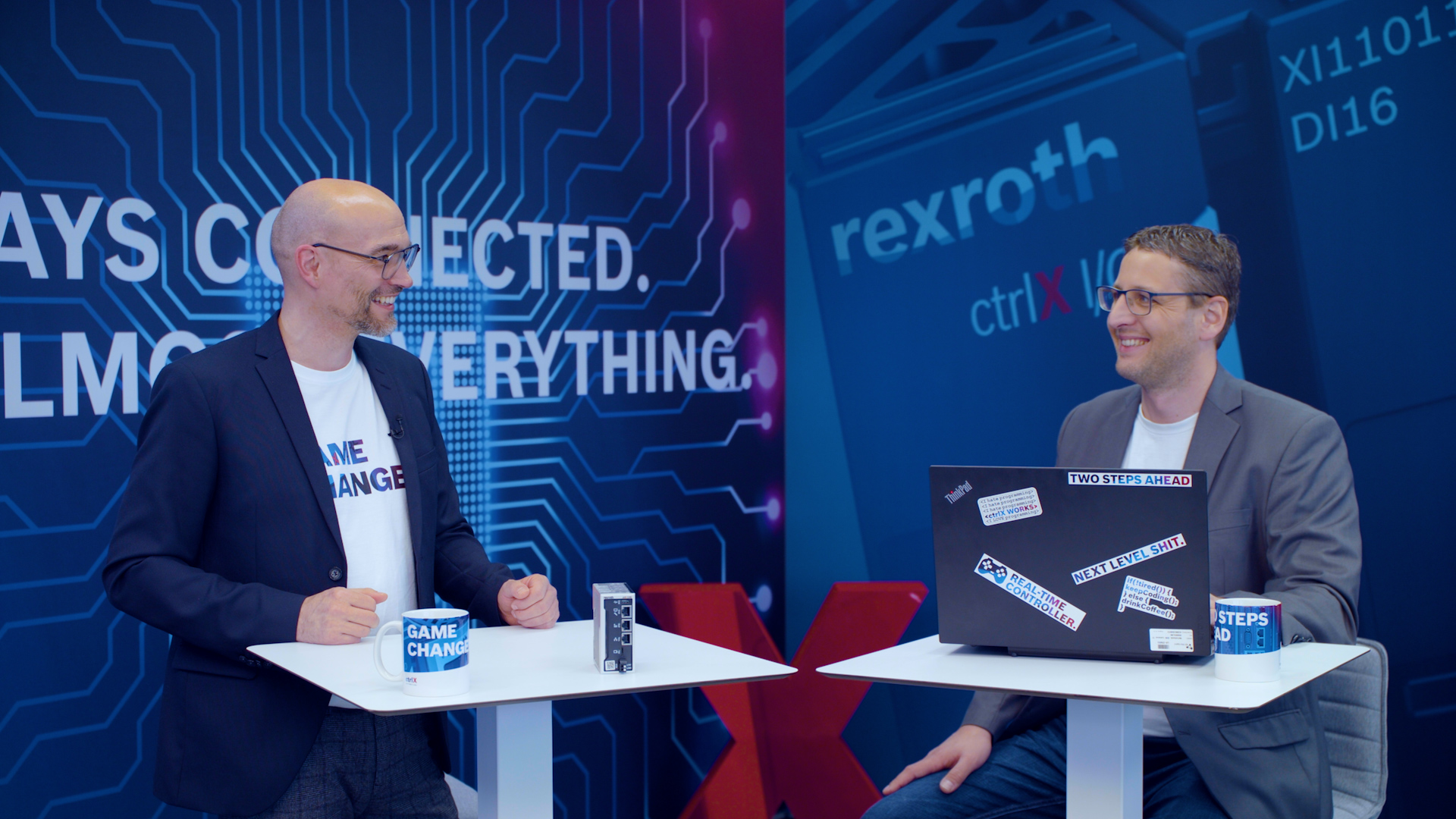 Gospodarz Christian Zentraf i jego gość Kai Müller stoją przy stołach konferencyjnych i opowiadają o konfiguracji aplikacji przeładunkowej za pomocą ctrlX CORE, sprzętu sterującego rozwiązania automatyzacji ctrlX AUTOMATION.